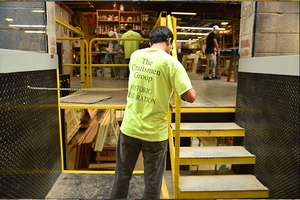 A man in a high-visibility yellow shirt that says The Craftsmen Group Historic Restoration stands near an industrial set of stairs leading into a cinderblock warehouse.