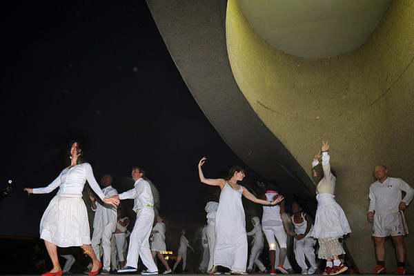 Young people wearing white dance at night near the curved support of a modern, concrete building.