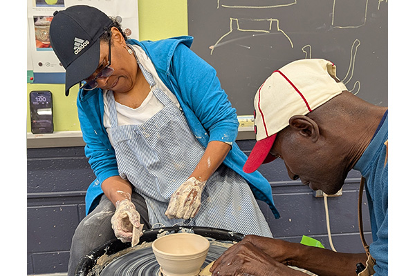 Two adults wearing aprons work over a pottery wheel on a small, clay bowl.
