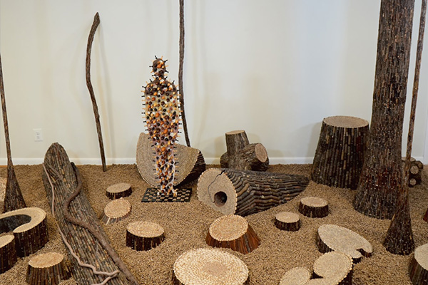 Art installation featuring sculptural, log-like forms surrounding a human figure covered in chess pieces.