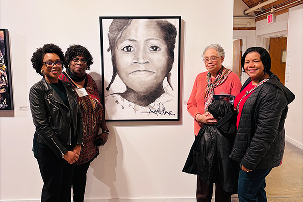 Four women pose next to a large, black-and-white portrait hanging on the wall of an art gallery.