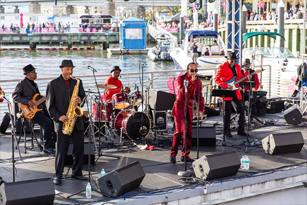 A band wearing red and black outfits performs on a stage at a waterfront dock.