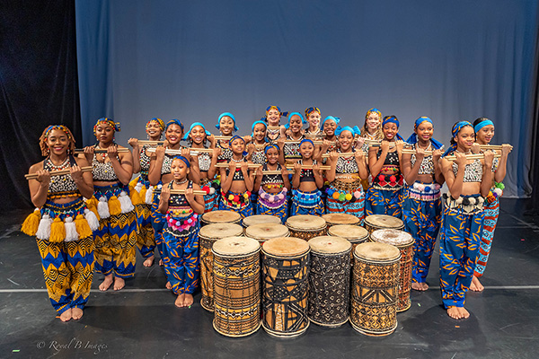 About 20 women in beaded tops and pants made of colorful, African print fabric pose around a collection of drums, all holding drumsticks in their hands.