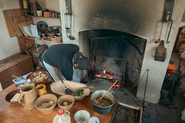 A person tends the fire in a large cooking hearth inside an 18th-century kitchen. Nearby is a table laden with crockery, pots, and bowls of food.