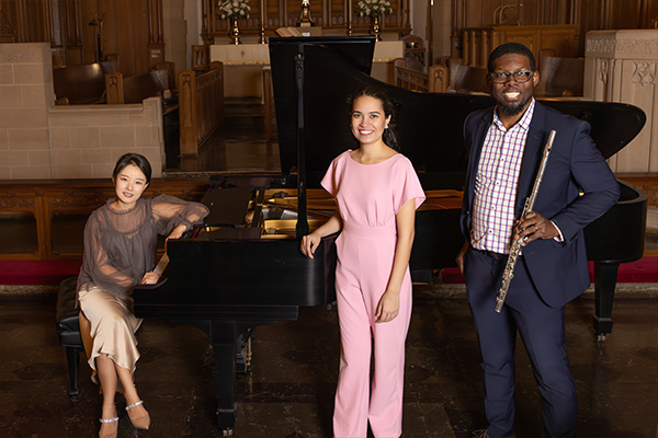 Three smiling musicians pose around a grand piano. One holds a flute.