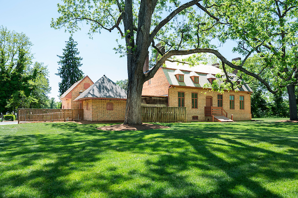 Historic brick house beside a large shade tree and expansive green lawn on a sunny day.