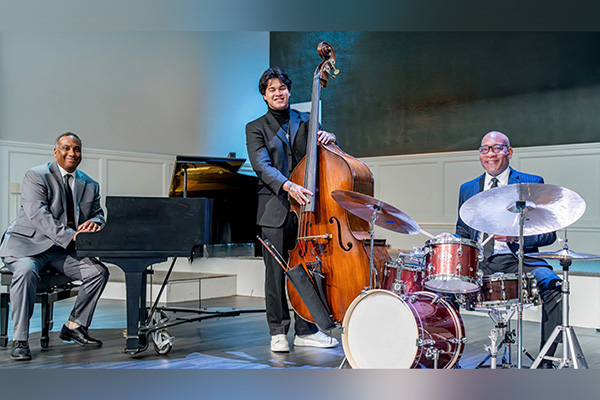Three smiling men in suits pose, smiling, on a stage with their musical instruments: a grand piano, an upright bass, and a drum kit.