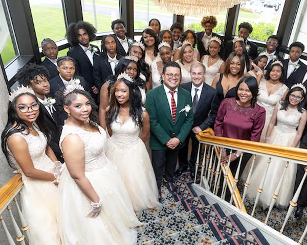 Several dozen people in formalwear pose on the landing of a large stairwell under a chandelier. The young women wear white gowns and tiaras; the young men wear tuxedos.
