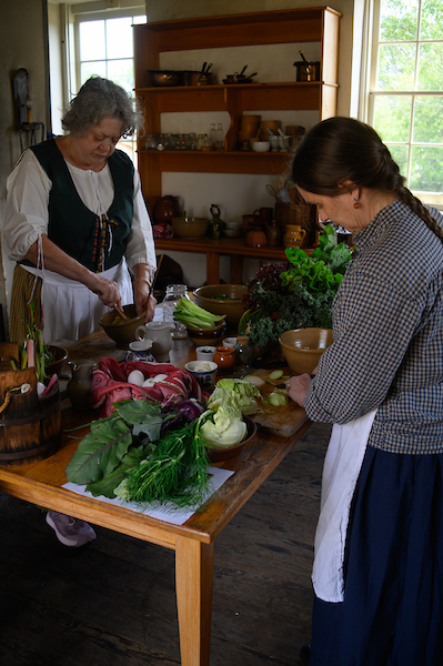 Two chefs prepare fresh vegetables at a wooden kitchen table inside a period-style historic home.