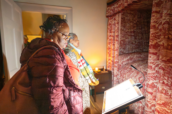 Visitors read an exhibit display inside a historic bedroom during a candlelit house tour.