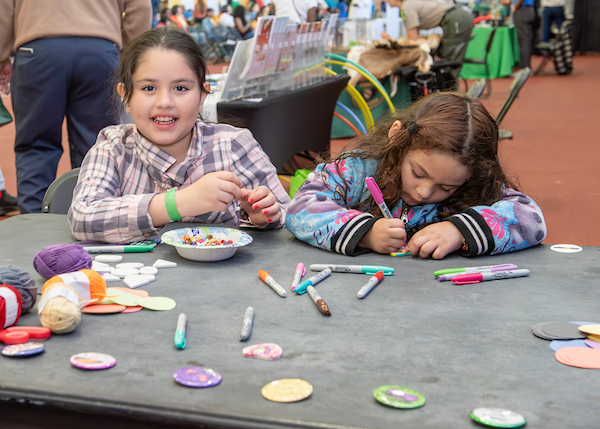 Two young girls sitting at a craft table filled with markers, yarn, and colorful buttons.